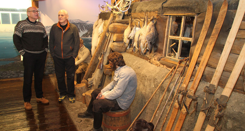 To menn står i en museumsutstilling med fangsthytte og fangstmann sittende utenfor